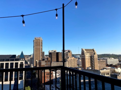 City skyline view from a balcony, featuring buildings and a clear blue sky with hanging lights.