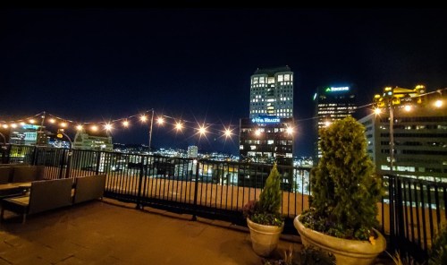 City skyline at night with illuminated buildings and string lights, viewed from a rooftop terrace.