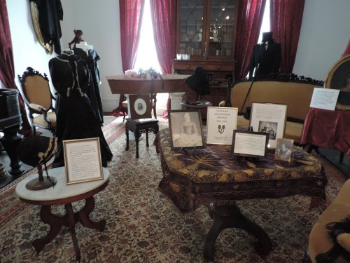 A vintage room with antique furniture, dresses, and framed photos, showcasing historical clothing and artifacts.