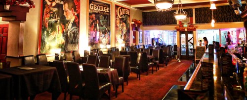 A cozy restaurant interior with dark chairs, tables, and vintage posters on the walls, illuminated by warm lighting.