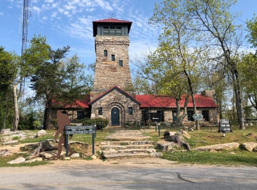 A stone building with a red roof and a tall tower, surrounded by trees and a pathway leading to the entrance.