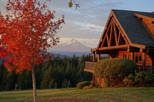 A log cabin with a mountain backdrop, surrounded by vibrant autumn foliage and a clear sky at sunset.