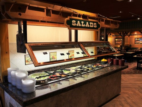 A salad bar with various fresh ingredients, bowls, and utensils in a restaurant setting.