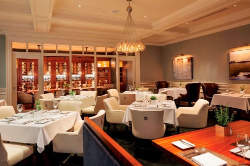 Elegant restaurant interior with white tablecloths, plush seating, and a wine display in a cozy, well-lit atmosphere.
