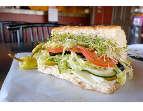 A fresh sandwich with layers of lettuce, tomato, cucumbers, olives, and sprouts, served with a pepper on the side.