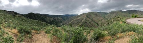 Panoramic view of rolling green hills and mountains under a cloudy sky, with a winding road in the foreground.