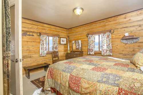 Cozy bedroom with wooden walls, a patterned quilt, a dresser, and a chair by the window. Warm, inviting atmosphere.