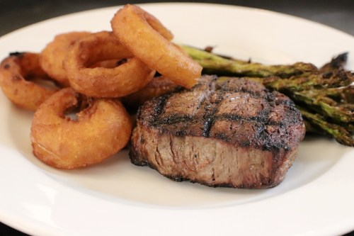 A plate featuring a grilled steak, crispy onion rings, and charred asparagus.