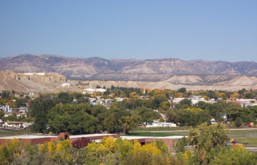 A scenic view of a town surrounded by mountains, with trees displaying autumn colors under a clear blue sky.