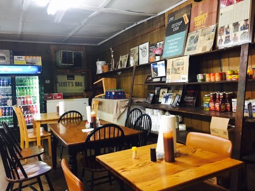 Interior of a rustic BBQ restaurant with wooden tables, shelves of sauces, and a drink cooler in the background.