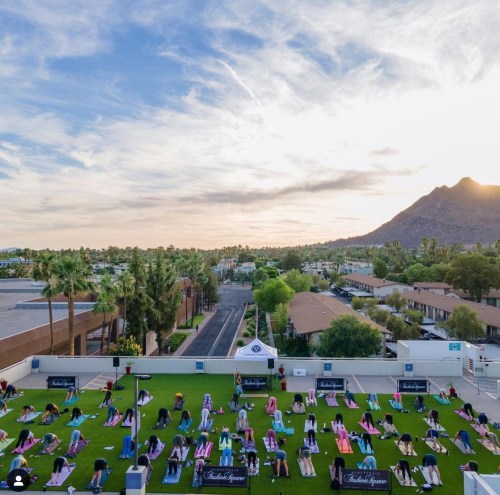 Aerial view of a yoga class on green grass, with participants practicing under a sunset and mountains in the background.