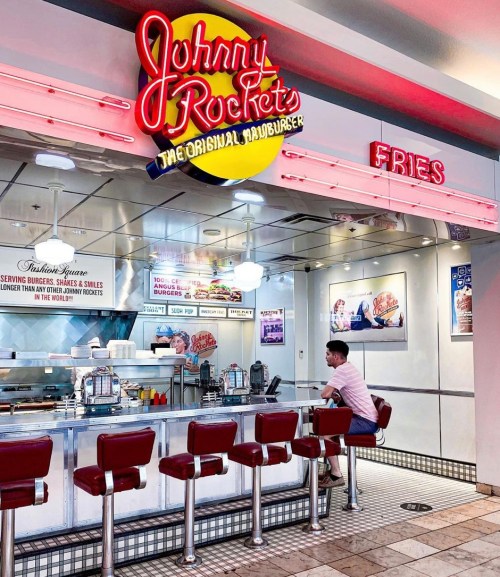 A diner-style restaurant with a neon sign reading "Johnny Rockets" and a customer sitting at the counter.