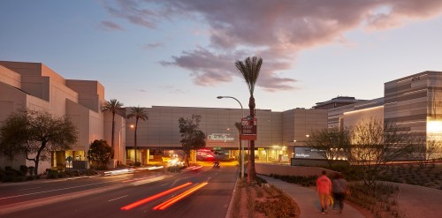 A modern shopping mall at dusk, with palm trees and light trails from passing cars on the street.