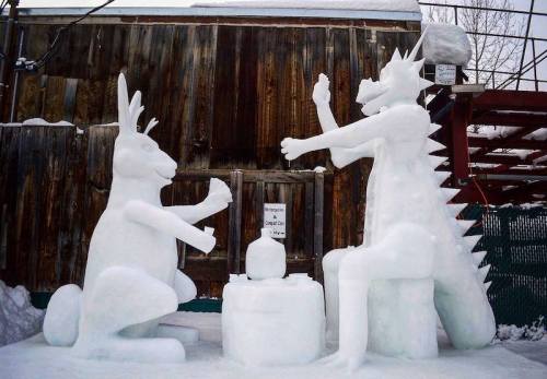 Two large snow sculptures: a rabbit and a dragon-like creature, sitting at a table in a snowy setting.