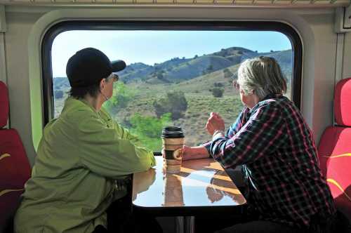 Two people sit at a table in a train, looking out the window at green hills and trees. A coffee cup is on the table.