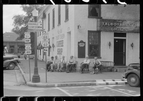 A group of men sit outside a coffee shop, with vintage signs and a historic building in the background.