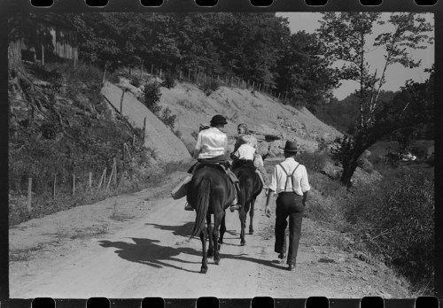 Three people riding horses along a dirt road, surrounded by trees and a hillside. Black and white photo.