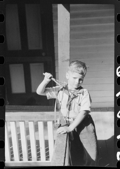 A young boy stands on a porch, holding a tool and looking at the camera with a serious expression.