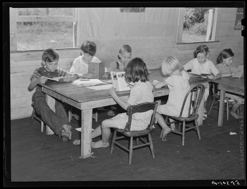 Children reading at a wooden table in a classroom, with sunlight streaming through the windows.