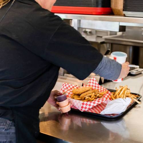 A person in a black shirt serves a tray with fried chicken, fries, and dipping sauces in a restaurant kitchen.