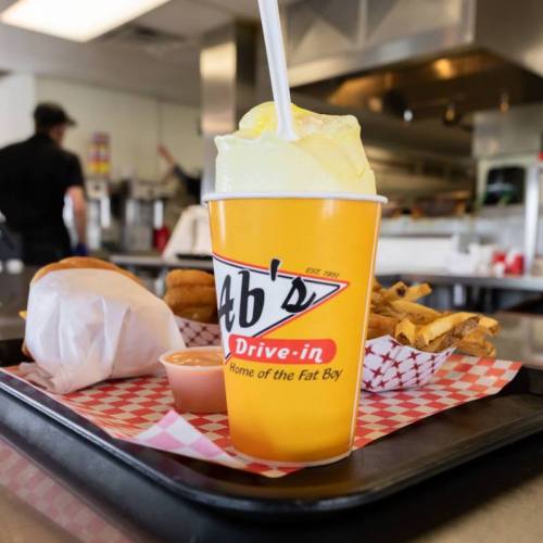 A yellow cup with a straw sits on a tray with fries, a burger, and dipping sauce at a drive-in restaurant.