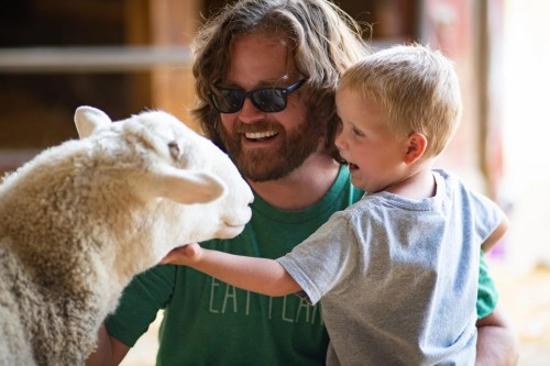 A man and a young boy smile while interacting with a sheep in a farm setting.