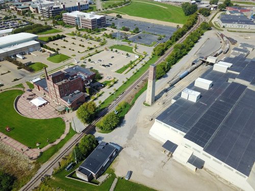 Aerial view of a park, buildings, and train tracks in an industrial area with green spaces and parking lots.