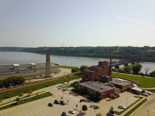 Aerial view of a river with a bridge, green trees, and a red brick building near a parking lot. Clear blue sky above.