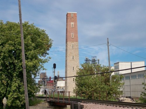 A tall brick and stone smokestack stands near industrial buildings and trees under a blue sky with scattered clouds.