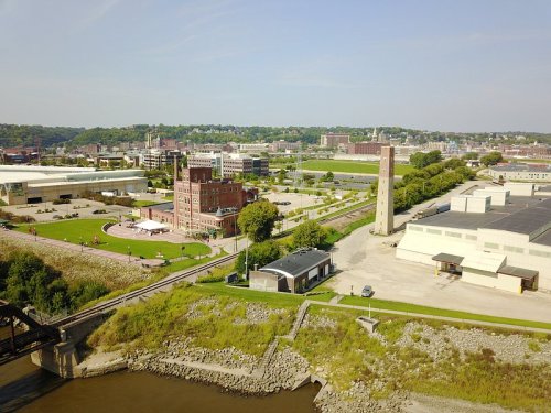 Aerial view of a riverside area featuring buildings, green spaces, and a clock tower in a city landscape.