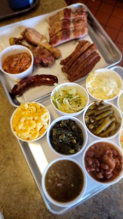A tray of barbecue featuring various meats and sides, including macaroni and cheese, greens, and beans in small cups.