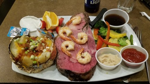 A plate featuring prime rib, shrimp, baked potato, mixed vegetables, and dipping sauces, with fruit on the side.