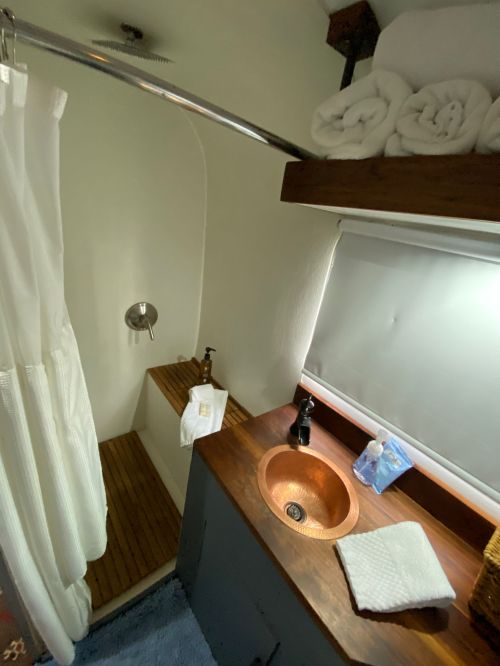 A small bathroom featuring a shower, wooden bench, sink with a copper faucet, and neatly stacked towels.