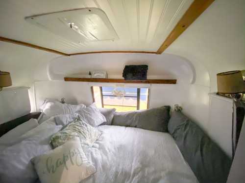 Cozy interior of a camper with a large bed, pillows, and a window showing a view outside. Warm, inviting atmosphere.