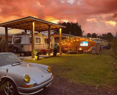 A vintage car parked in front of a cozy outdoor space with a trailer, under a vibrant sunset sky.