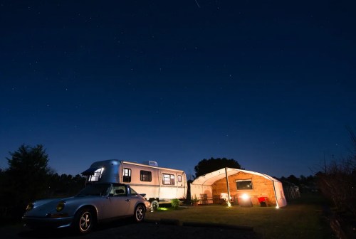 A silver camper and a vintage car are parked under a starry night sky, with a cozy tent nearby.