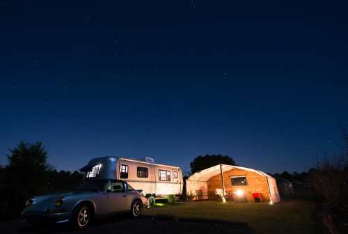 A silver camper and a vintage car are parked under a starry night sky, with a cozy tent nearby.