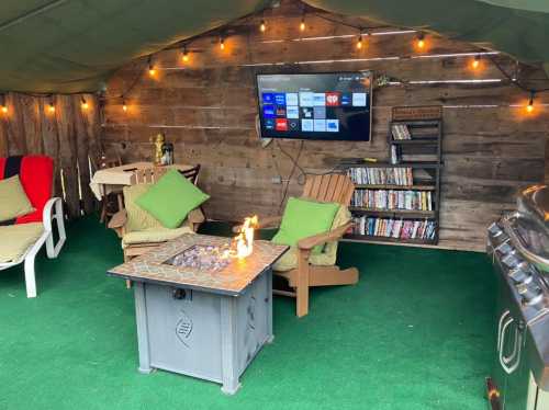 Cozy outdoor space with two chairs, a fire pit table, a TV, and a bookshelf, all under warm string lights.