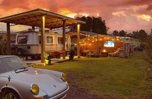 A vintage silver car parked in front of a cozy outdoor space with a trailer, string lights, and a sunset sky.