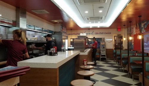 A bright diner interior with staff preparing food, checkered floor, and retro decor featuring "Baltimore Legend Since 1943."