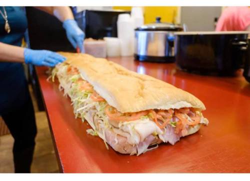 A person in blue gloves prepares a long sub sandwich loaded with lettuce, tomatoes, and turkey on a red countertop.