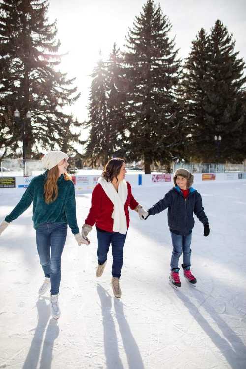 Three people ice skating together, holding hands, with trees and sunlight in the background.