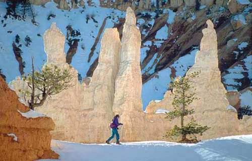 A person walks through a snowy landscape with towering rock formations and pine trees in the background.