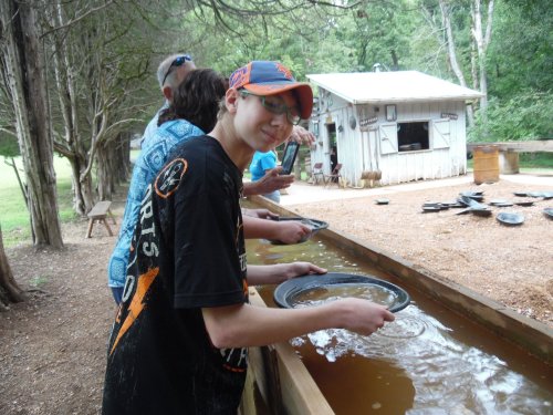 A young boy pans for gold in a shallow stream, surrounded by others at a rustic outdoor setting.