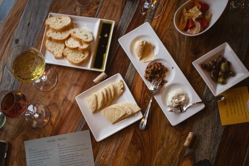 A wooden table with plates of bread, cheese, olives, and a wine menu, accompanied by glasses of wine.