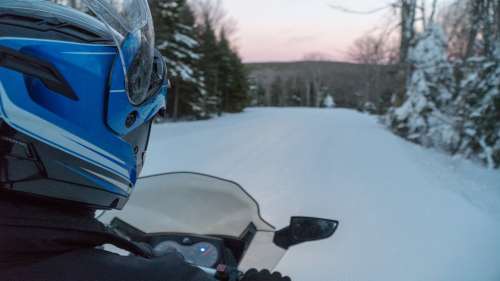 A person on a snowmobile rides down a snowy trail surrounded by trees at dusk.