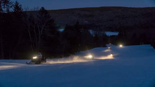Snowmobiles ride through a snowy landscape at dusk, with headlights illuminating the path ahead.