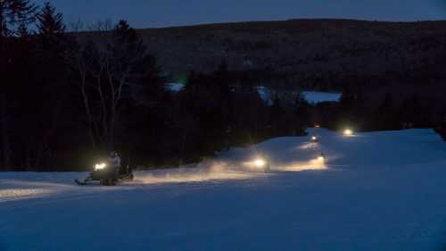 Snowmobiles ride through a snowy landscape at dusk, with headlights illuminating the path ahead.