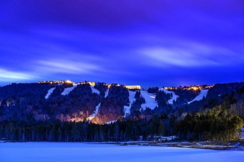 A serene winter landscape at dusk, featuring a snowy hill with lights and a calm lake reflecting the twilight sky.