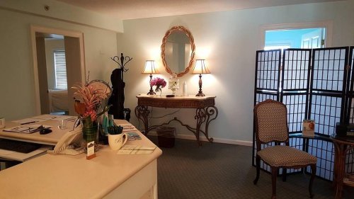 A cozy reception area with a desk, decorative mirror, lamps, and a chair, featuring soft blue walls and floral accents.